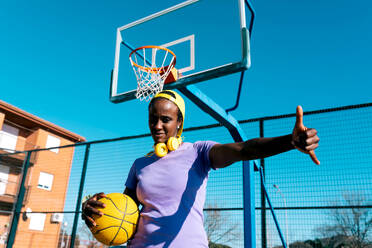 Low angle of cool young African American female athlete in sportswear and cap with basketball and headphones showing shaka sign and looking at camera on sports ground in sunlight - ADSF45434