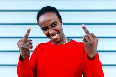 Portrait of naughty black female model looking at camera and showing middle fingers while standing against blue wall on street - ADSF45446