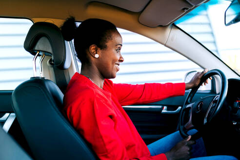 Side view of positive adult African American woman with dark hair in red sweater smiling while sitting in modern car on sunny day - ADSF45453