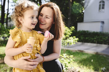 Lächelnde Mutter betrachtet ihre Tochter, die einen Seifenblasenstab hält - EYAF02707