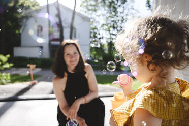 Mutter betrachtet ihre Tochter beim Blasen von Seifenblasen an einem sonnigen Tag - EYAF02709