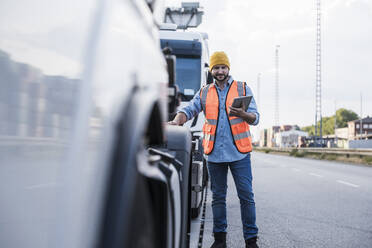 Smiling truck driver wearing knit hat standing with tablet PC by truck - UUF29569