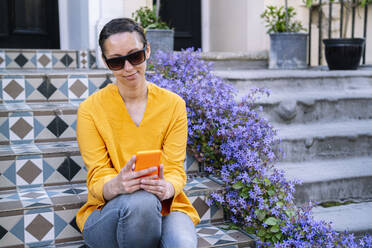 Frau mit Sonnenbrille und Smartphone auf einer Treppe sitzend - AMWF01563