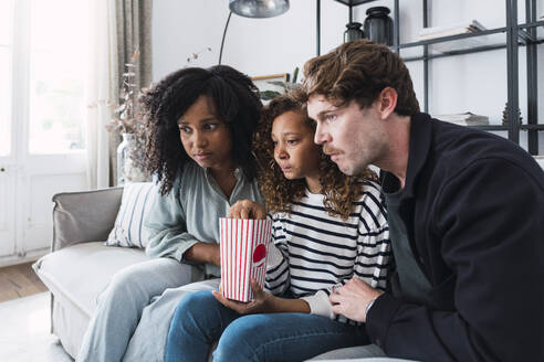 Eltern und Tochter sitzen auf dem Sofa, essen Popcorn und schauen einen Film auf dem Laptop - PNAF05617