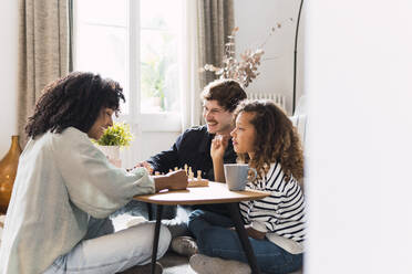 Eltern sitzen im Wohnzimmer und bringen ihrer Tochter das Schachspielen bei - PNAF05642