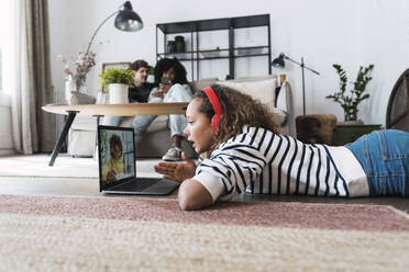 Mädchen mit Videoanruf mit Freund auf dem Boden liegend, während die Eltern auf der Couch im Hintergrund sitzen - PNAF05680