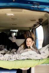 Thoughtful young traveler with her hand on her chin lying on a soft blanket and looking away while traveling in a camper van - ADSF45454