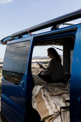 Side view of unrecognizable woman traveler messaging on her smartphone while sitting in a blue camper van parked in a sunny lot during a summer trip - ADSF45455