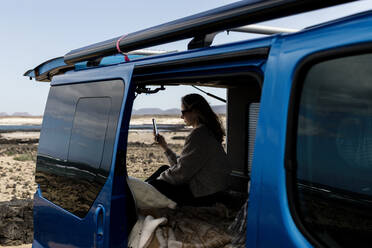 Side view of unrecognizable young traveler messaging on her smartphone while sitting in a blue camper van parked in a sunny lot during a summer trip - ADSF45456