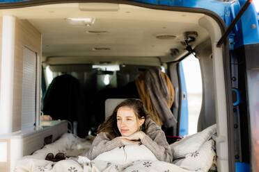 Pensive young female traveler in casual outfit lying on soft blanket and looking away while traveling in camper van during summer holidays - ADSF45458