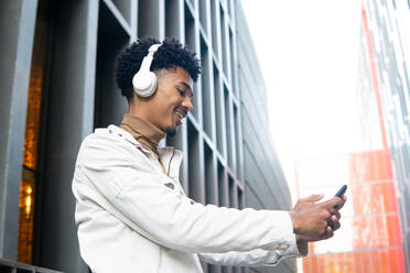 Ein lächelnder junger afroamerikanischer Mann mit drahtlosen Kopfhörern und Afrofrisur macht ein Selfie, während er seine Musik genießt - ADSF45464