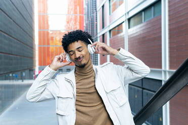 Positive African American young with wireless headphones eyes closed and curly hair listening to music while walking on a city street - ADSF45465