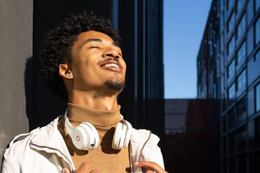 Cheerful African American young with afro hairstyle and closed eyes leaning against a wall with headphones around his neck - ADSF45466