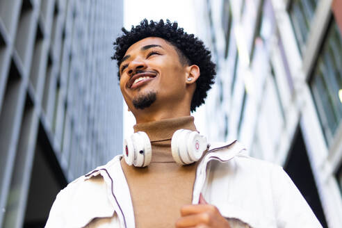 From below a smiling young African American man with afro hair looking away with headphones around his neck in front of modern buildings - ADSF45468