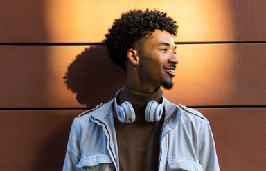 Happy African American young man with afro hairdo and headphones around his neck smiling and looking away from a wall on a street in the sunlight - ADSF45469