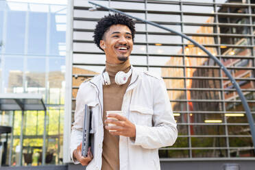 Happy African American young man with headphones around his neck and laptop computer looking away as he walks against a modern building - ADSF45472