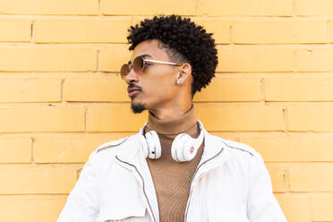 Serious young African American man with sunglasses afro hairstyle and headphones leaning against a brick wall in a sunny street - ADSF45475