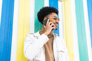 Positive African American young with sunglasses combed afro talking on his mobile phone and smiling while looking away against a wall - ADSF45478