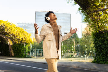 Happy African American young with eyes closed listening to music with headphones and arms open as he walks down the street against the trees - ADSF45479