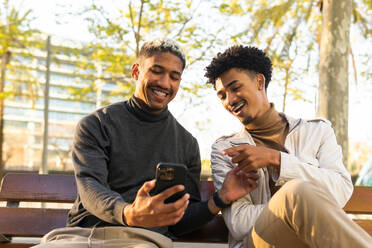 Low angle of happy young African American male friends smiling and browsing social media on their smartphone while sitting on a wooden bench - ADSF45480