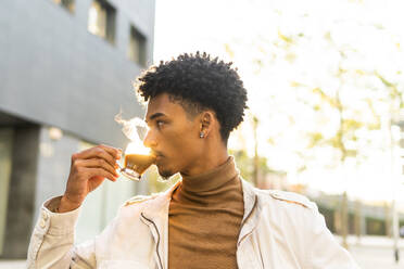 Thoughtful young African American male in casual wear with Afro hairstyle drinking hot coffee from transparent cup while looking away on sunny street - ADSF45481