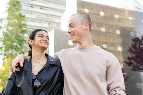 Low angle of smiling stylish young boyfriend and girlfriend in casual clothes hugging on city street while looking at each other - ADSF45484