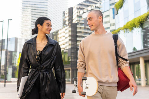 Positive young girlfriend in leather coat an boyfriend in beige outfit with beige outfit walking on city street and looking at each other - ADSF45485