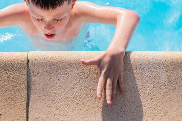 High angle of kid with closed eyes in swimming pool water with opened mouth near edge of poolside with hand on hot concrete platform during summertime - ADSF45500