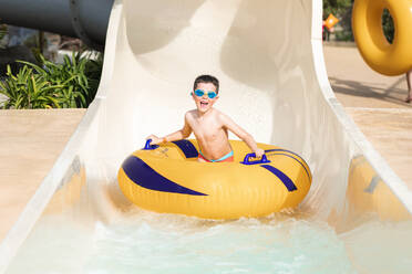 Happy shirtless boy in swimming trunks and goggles holding an inflatable ring at the exit of a slide and looking at the camera against the sunlight - ADSF45503