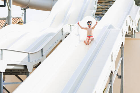 Low angle of a happy child with swimming goggles on open arms in a swimsuit sliding down a slide having fun while looking at the camera in a park - ADSF45506