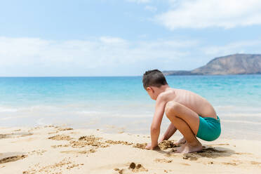 Side view of shirtless boy sitting on sandy beach near sea and mountain ridge while having fun during sunny day - ADSF45507