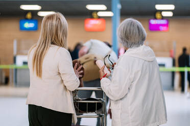 Back view of unrecognizable calm elderly female travelers in casual wear with baggage in airport terminal together while departing for vacation - ADSF45515