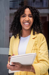 Cheerful young African American girl in yellow jacket and curly hair looking at the camera and writing notes on a notepad while smiling - ADSF45519