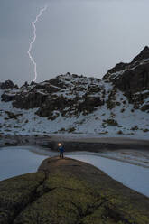 Back view of unrecognizable traveler with bright lamp standing on edge of rock opposite river and snowy mountains with lightning sky in winter - ADSF45525