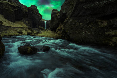 Picturesque view of waterfall flowing through rocky mountains near river under green aurora borealis in Iceland - ADSF45532