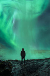 Back view of traveler standing admiring green majestic polar lights during trip through Iceland mountains - ADSF45533