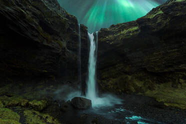 Picturesque view of waterfall flowing through rocky mountains near river under green aurora borealis in Iceland - ADSF45535