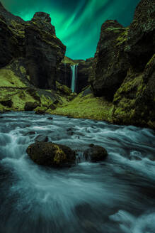 Picturesque view of waterfall flowing through rocky mountains near river under green aurora borealis in Iceland - ADSF45537