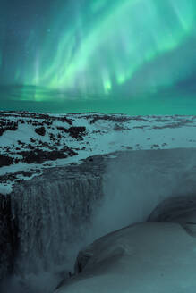 Picturesque scenery of powerful waterfall falling from snowy slope under green aurora borealis illuminating night sky in Iceland - ADSF45539