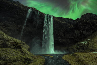 Picturesque scenery of waterfall flowing through mountains under green aurora borealis at night in Iceland - ADSF45541