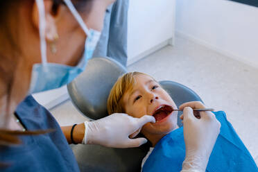 High angle of cropped unrecognizable female dentist in uniform and gloves with mask using dental tool to examine teeth of child in dental clinic - ADSF45556