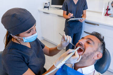 Professional dentist in medical mask and latex gloves examining teeth of male patient with open mouth and using table in modern dental clinic - ADSF45557