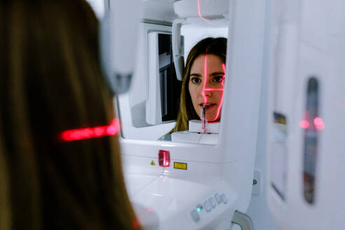 Young female in casual clothes standing and using dental x ray digital scanner panorama machine in bright clinic - ADSF45558