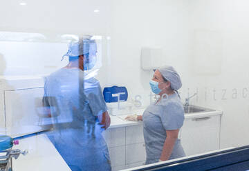 Side view of female dentist in medical uniform with back view of male colleague with protective mask standing and working in modern clinic - ADSF45559