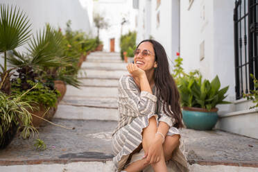 Positive female in sunglasses smiling looking away sitting with hand at chin on stairs on street with potted plants against blurred white buildings - ADSF45563