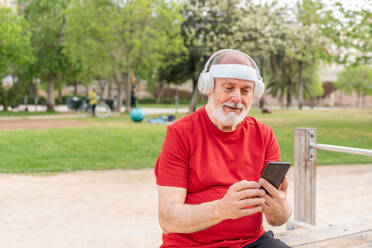 Positive senior bearded male in casual clothes and headphones browsing mobile phone while listening to music in wireless headphones in park - ADSF45581