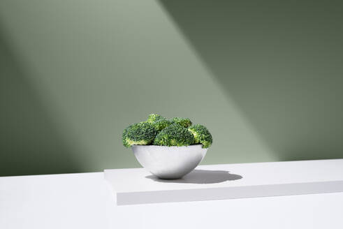 Fresh green broccoli growing in white bowl on plain table against gray background under bright ray of light - ADSF45585