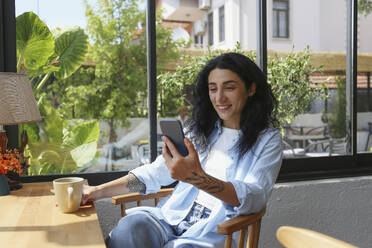 Lächelnde Frau, die ein Smartphone benutzt und auf einem Stuhl in einem Café sitzt - SYEF00510