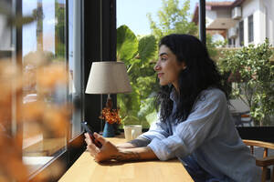 Lächelnde Frau schaut aus dem Fenster und sitzt in einem Café - SYEF00511