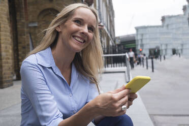 Lächelnde Geschäftsfrau sitzt mit Smartphone vor einem Gebäude - AMWF01569